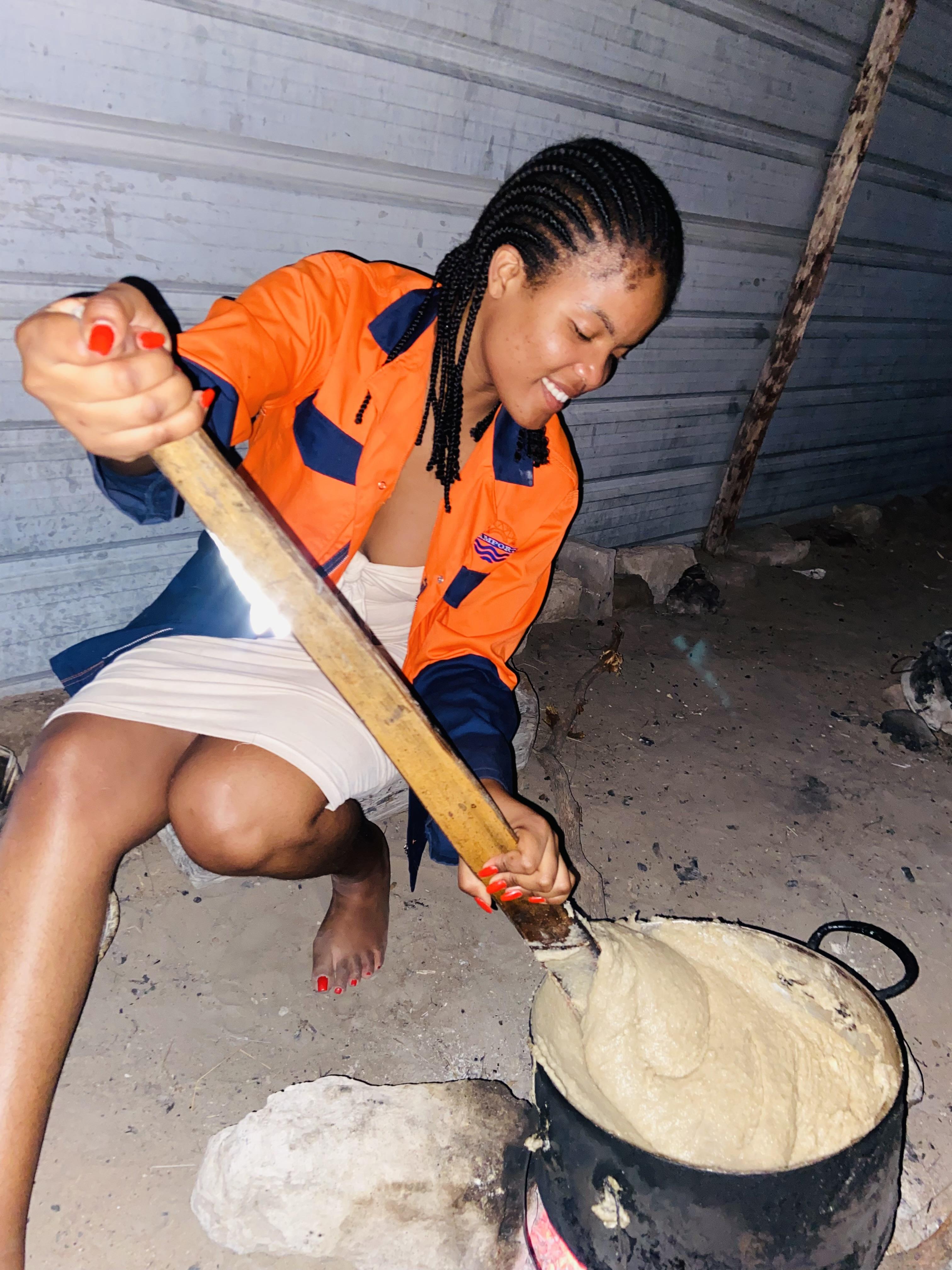 A cook stirring thick porridge in a black pot over a fire with a long wooden paddle
