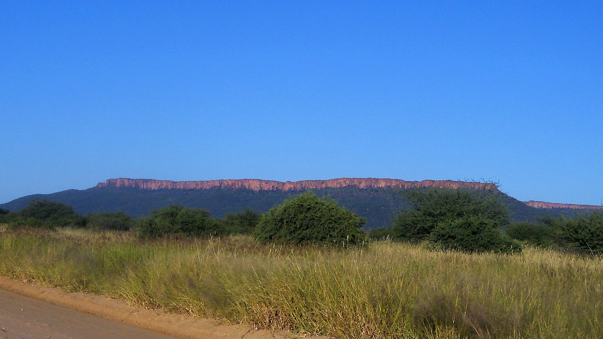 Red sandstone cliffs of the Waterberg Plateau above green bush, seen from a gravel road